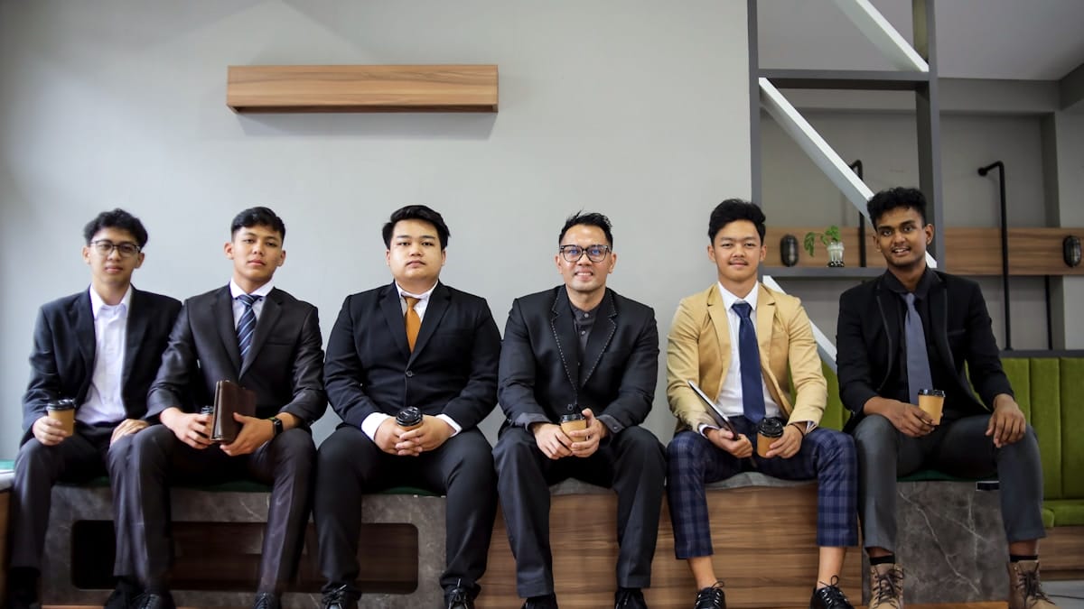 Six men in business attire sit on a bench indoors, each holding a drink, facing the camera and smiling slightly.