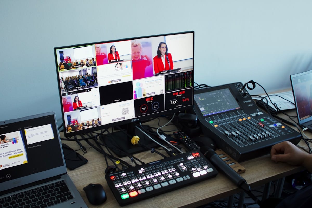 A video production workstation with a monitor showing a multi-camera feed, audio mixer, switcher, and various cables on a desk.