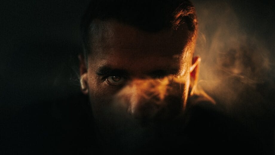 A close-up of a persons face partially obscured by dramatic lighting and smoky haze, with intense eyes visible.