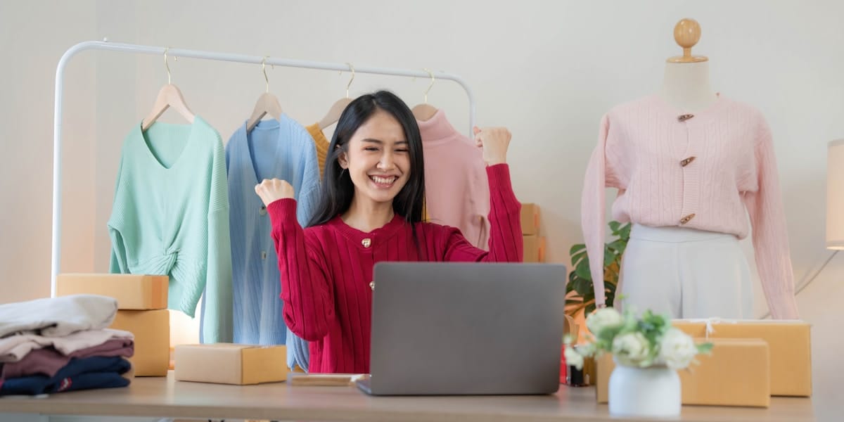 Woman sitting at a desk with a laptop, surrounded by clothing and packages, smiling with raised fists in a home office setting.