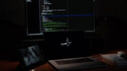 A laptop, tablet, and large monitor display code and images on a dimly lit desk with a book beside the laptop.