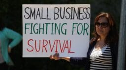 A woman holds a sign that reads, Small business fighting for survival, during a daytime outdoor protest.