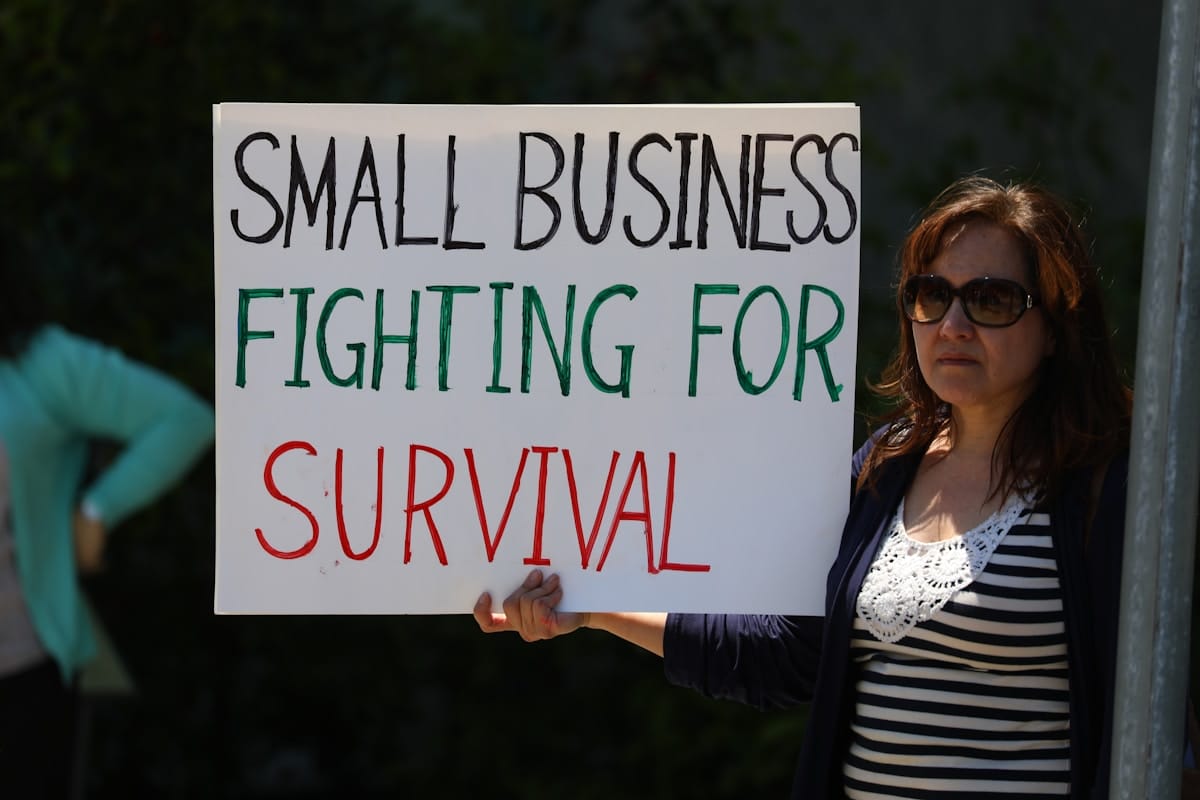 A woman holds a sign that reads, Small business fighting for survival, during a daytime outdoor protest.