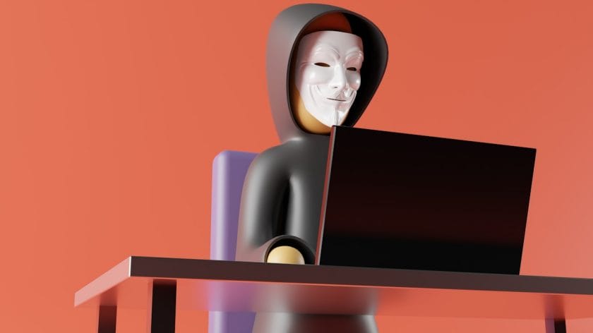A hacker in a hooded sweatshirt and white mask sits at a desk using a laptop against an orange background.