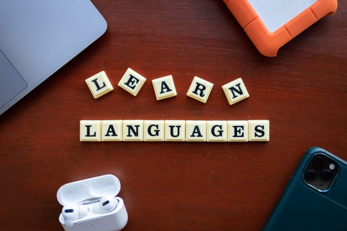 Scrabble tiles spelling “LEARN LANGUAGES” on a wooden table with a laptop, phone, orange hard drive, and earbuds nearby.