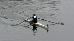A single rower wearing number 1 rows a scull boat on calm water, creating ripples behind them.