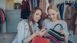 Two women sitting on a couch with shopping bags, looking at a smartphone and smiling in a clothing store.