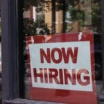 A red and white Now Hiring sign is displayed in the window of a storefront.