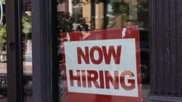 A red and white Now Hiring sign is displayed in the window of a storefront.