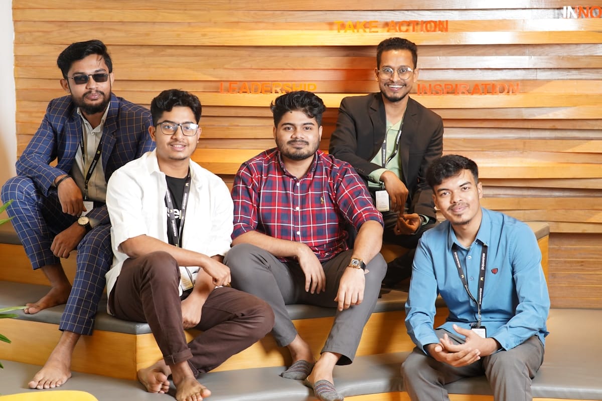 Five men sit and pose for a group photo on tiered wooden steps with motivational words on the wall behind them.