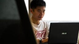 A person sits at a desk working on an ASUS laptop, wearing a white T-shirt, with natural light coming through a window.