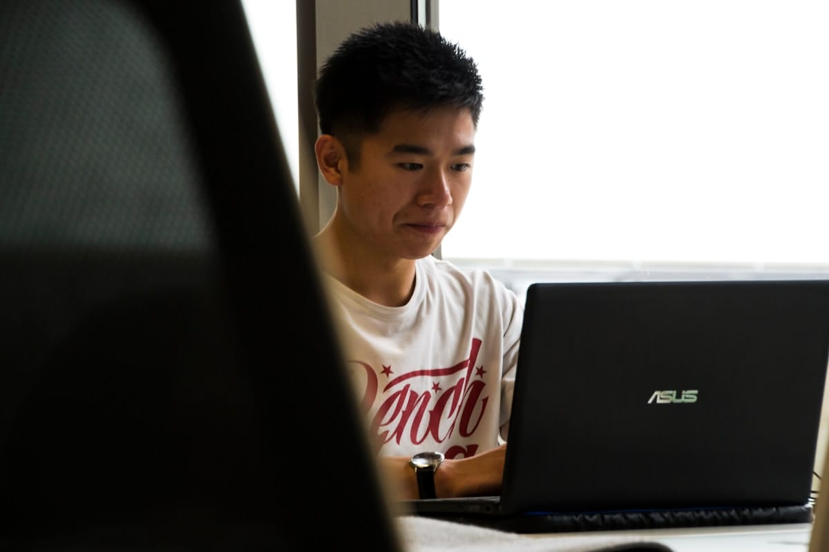 A person sits at a desk working on an ASUS laptop, wearing a white T-shirt, with natural light coming through a window.