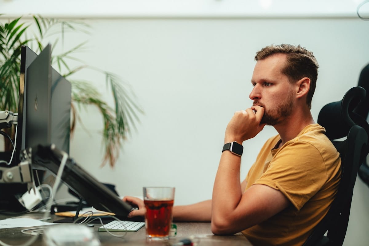 Man in a yellow shirt sits at a desk, looking at a computer monitor with a glass of tea in front of him.