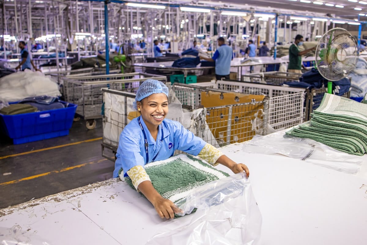A worker in a blue uniform and hairnet folds towels at a table in a brightly lit textile factory.