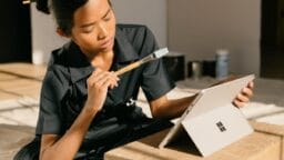 Person sitting on the floor, holding a paintbrush, and looking thoughtfully at a Microsoft Surface laptop on a wooden box.