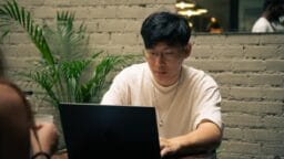 Person with glasses using a laptop at a table in a modern indoor space with a brick wall and plants.