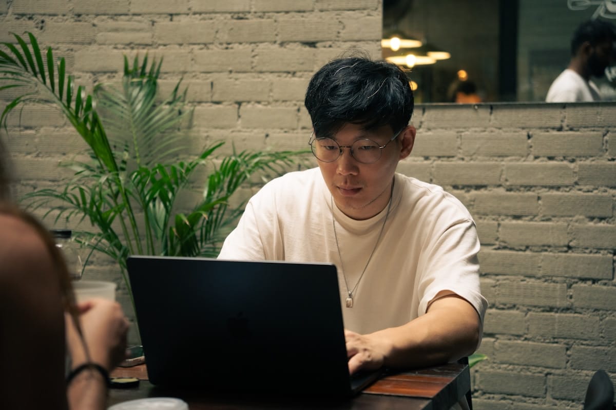 Person with glasses using a laptop at a table in a modern indoor space with a brick wall and plants.