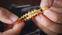 Two hands holding a detailed gold pendant shaped like a spiral shell or stylized corn cob.