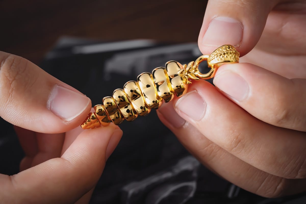 Two hands holding a detailed gold pendant shaped like a spiral shell or stylized corn cob.