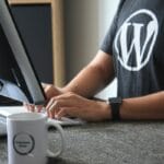 Person wearing a WordPress t-shirt types on a keyboard at a desk with a computer monitor and a mug labeled WordPress OKULLU.