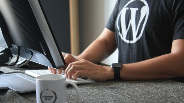 Person wearing a WordPress t-shirt types on a keyboard at a desk with a computer monitor and a mug labeled WordPress OKULLU.