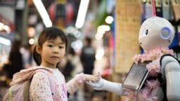 A young girl holds hands with a humanoid robot decorated with pink flowers in a busy market setting.