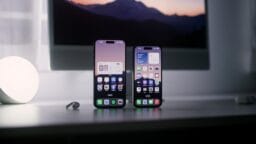 Two smartphones stand upright on a desk, displaying their home screens, with a computer monitor and accessories in the background.