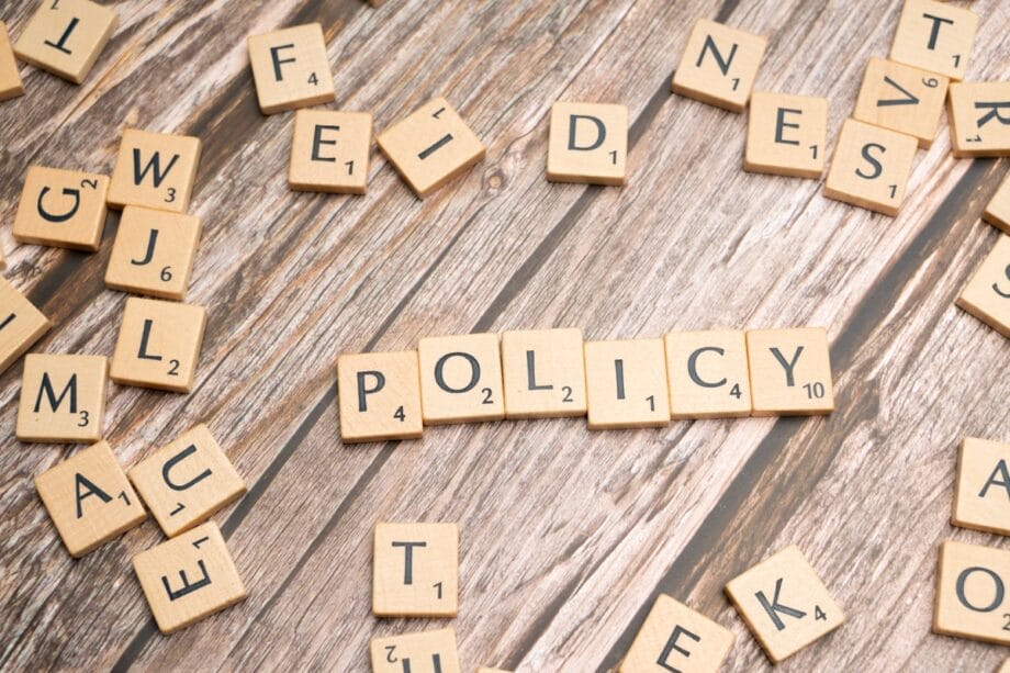 Scrabble tiles spelling POLICY on a wooden surface, surrounded by scattered letter tiles.