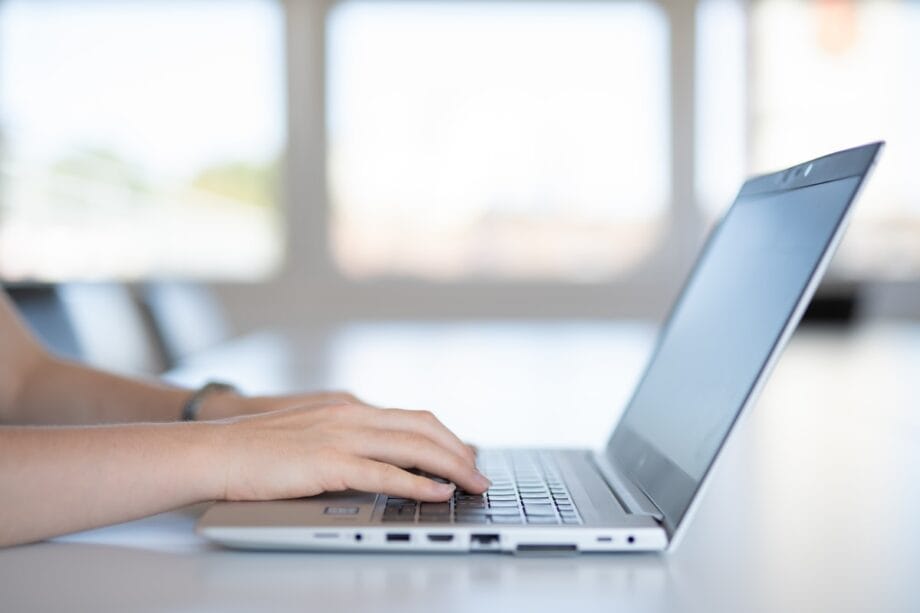 Person typing on a silver laptop at a desk in a bright room with large windows in the background.