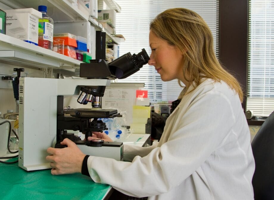 A person in a lab coat looks through a microscope in a laboratory with various scientific equipment and supplies.