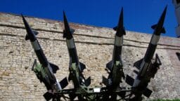 Four surface-to-air missiles are mounted on a military launcher in front of a stone wall under a clear blue sky.