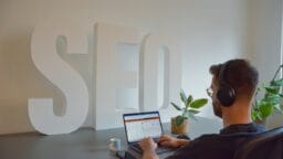 Person wearing headphones works on a laptop at a desk with large white SEO letters and a potted plant in the background.