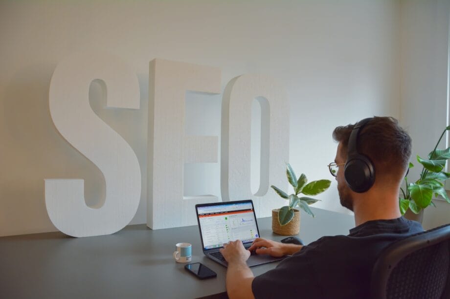 Person wearing headphones works on a laptop at a desk with large white SEO letters and a potted plant in the background.