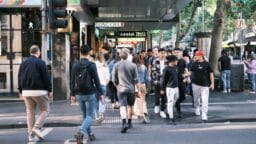 People walk across a busy city street intersection with shops, signs, and trees visible in the background.