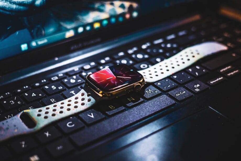 A smartwatch with a white strap rests on the keyboard of a black laptop, with a screen visible in the background.