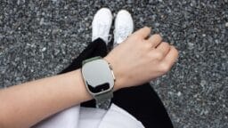 A person wearing a smartwatch with a green strap stands on gravel, visible from above with white shoes and black pants.