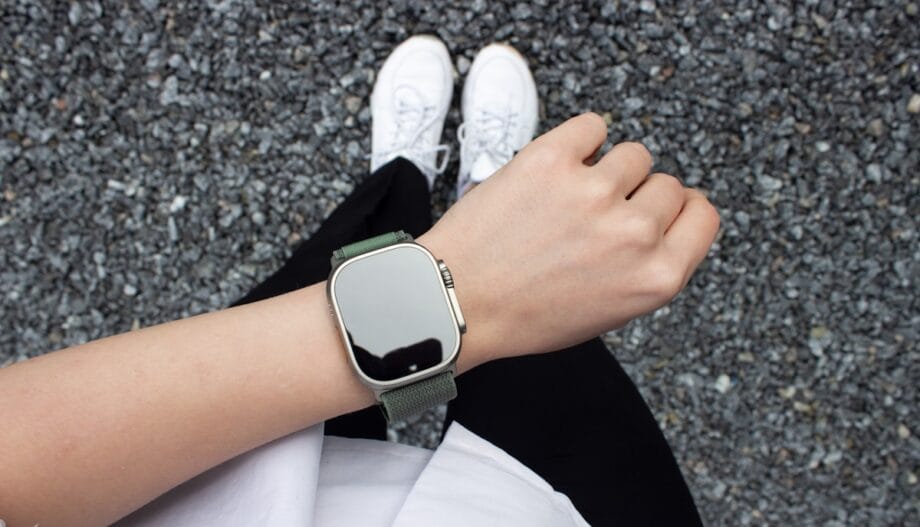 A person wearing a smartwatch with a green strap stands on gravel, visible from above with white shoes and black pants.