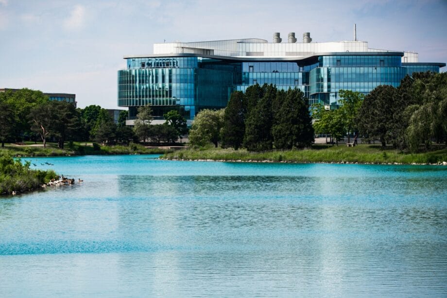 Modern glass office building with trees in front, reflected in a calm blue lake under a partly cloudy sky.