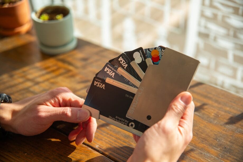 A person holds a metal wallet containing several credit and debit cards, fanned out on a wooden table in sunlight.