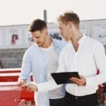 Two men examine red machinery outdoors; one holds a clipboard and pen while discussing details with the other.
