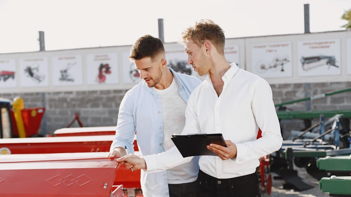 Two men discussing agricultural equipment purchase at a dealership, one holding a clipboard outside.
