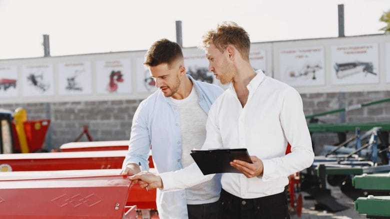 Two men examine red machinery outdoors; one holds a clipboard and pen while discussing details with the other.