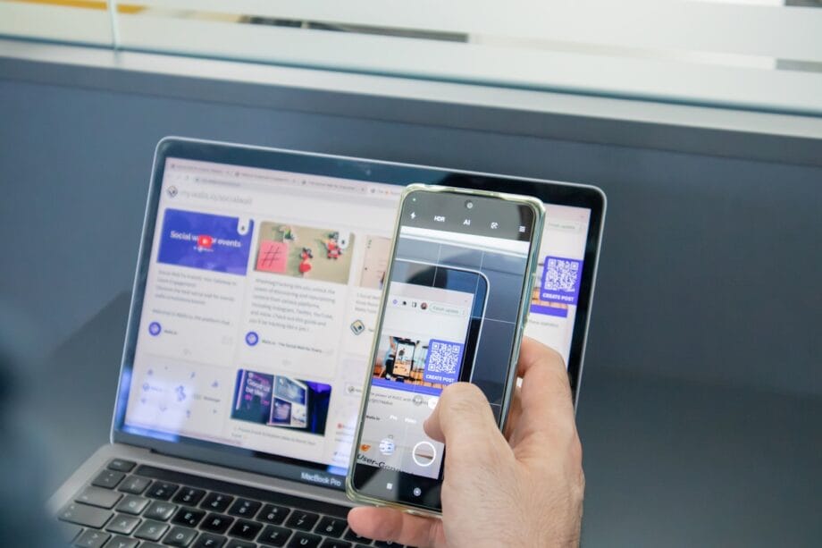 Person holding a smartphone to scan a QR code displayed on a laptop screen showing a website with social media content.