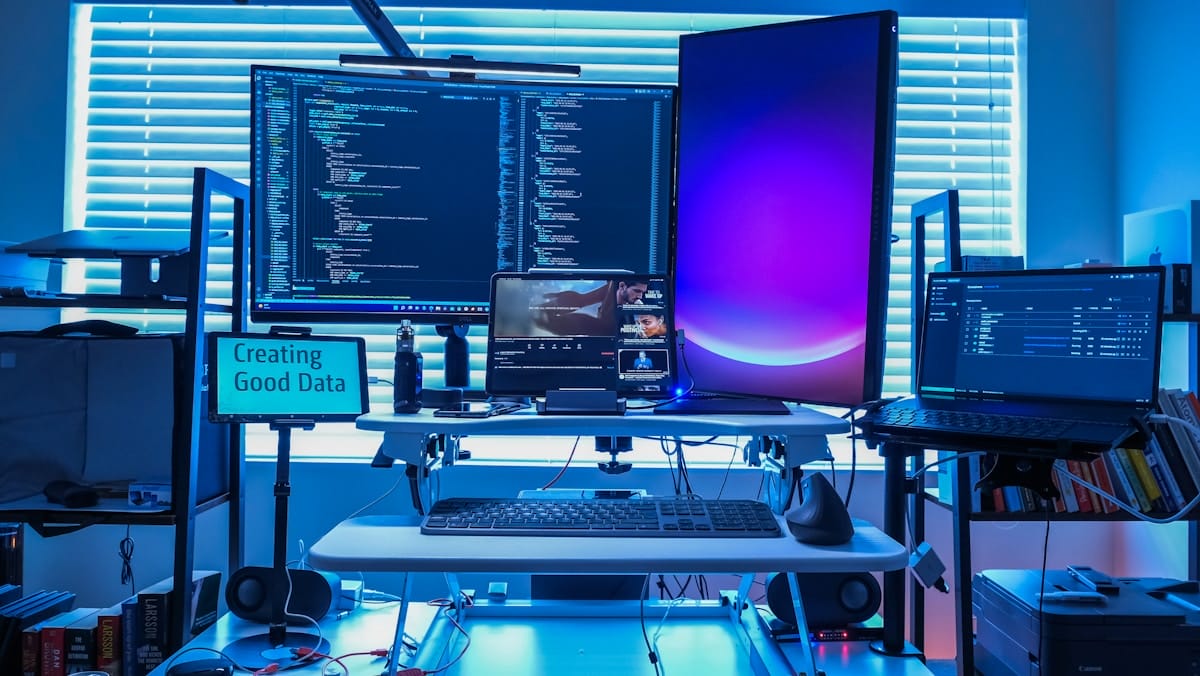A modern desk setup with multiple monitors, a laptop, a keyboard, and various tech devices in a blue-lit room.