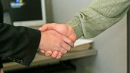 Two people shaking hands in an office setting, with a computer monitor visible in the background.