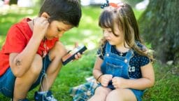 Two young children sit on grass under a tree, sharing earphones and listening to audio from a smartphone.