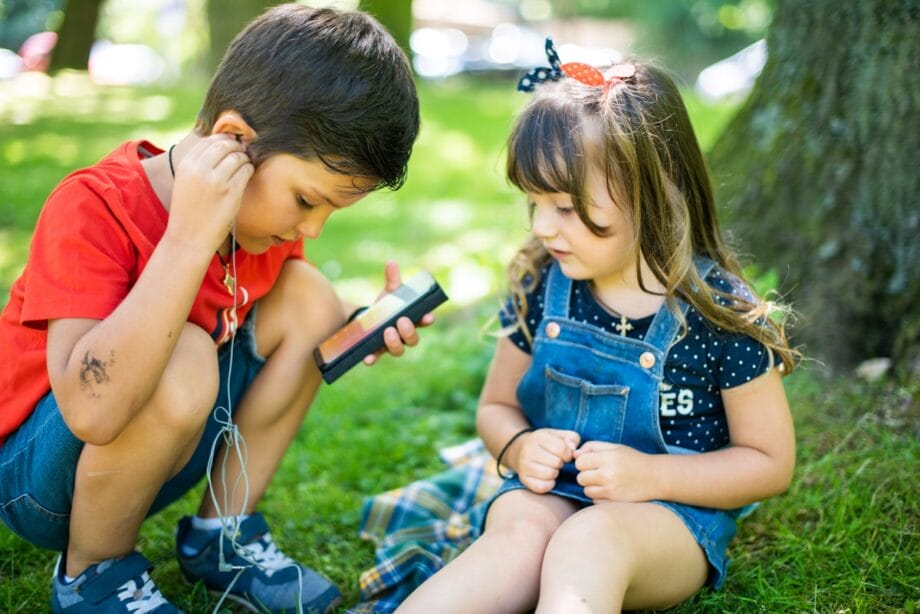 Two young children sit on grass under a tree, sharing earphones and listening to audio from a smartphone.