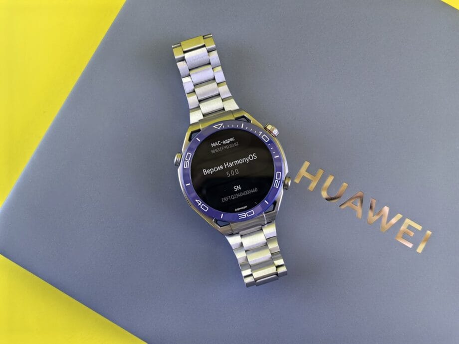 A Huawei smartwatch displaying HarmonyOS 5.0 is placed on a closed Huawei laptop with a yellow background.