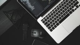 Laptop displaying code next to a smart watch, wireless mouse, and phone on a black desk.