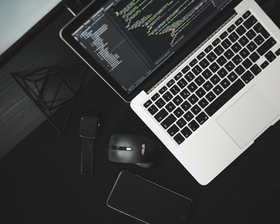 Laptop displaying code next to a smart watch, wireless mouse, and phone on a black desk.